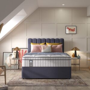 A full shot of the Spencer Plush Mattress, shown on a dark blue divan base with a tall, contemporary upholstered headboard, set in a bright bedroom with light wood wall panelling. The mattress has a deep quilted white top.
