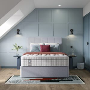 A full shot of the Blake Medium Mattress, shown on a light grey divan base with a matching upholstered headboard, set in a contemporary bedroom with light blue wall panelling. The mattress has a deep quilted white top.