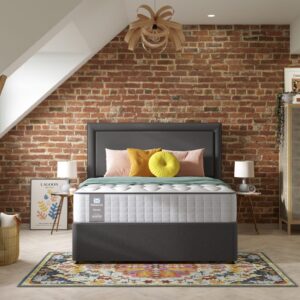 A full shot of the Emin Firm Mattress, shown on a dark grey divan base with a matching headboard, set in a contemporary bedroom with a brick feature wall. The mattress has a classic quilted white top.
