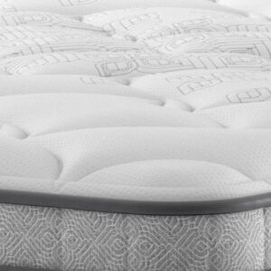 A full shot of the Spencer Plush Mattress, shown on a dark blue divan base with a tall, contemporary upholstered headboard, set in a bright bedroom with light wood wall panelling. The mattress has a deep quilted white top.