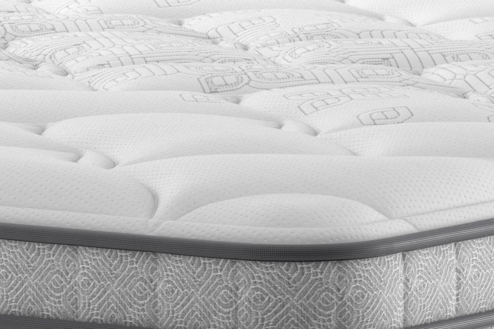 A full shot of the Spencer Plush Mattress, shown on a dark blue divan base with a tall, contemporary upholstered headboard, set in a bright bedroom with light wood wall panelling. The mattress has a deep quilted white top.