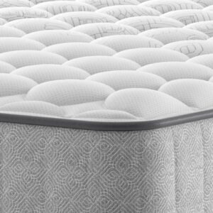 A full shot of the Emin Firm Mattress, shown on a dark grey divan base with a matching headboard, set in a contemporary bedroom with a brick feature wall. The mattress has a classic quilted white top.