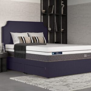 A full shot of the Phoenix 2900 Hybrid Mattress, shown on a deep blue divan base with a contemporary upholstered headboard, set in a modern bedroom with dark shelving. The mattress features a white top panel and a distinctive grey quilted side panel.
