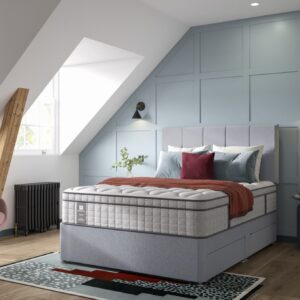 A full shot of the Blake Medium Mattress, shown on a light grey divan base with a matching upholstered headboard, set in a contemporary bedroom with light blue wall panelling. The mattress has a deep quilted white top.