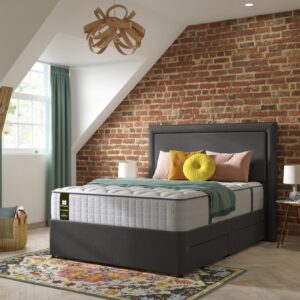 A full shot of the Emin Firm Mattress, shown on a dark grey divan base with a matching headboard, set in a contemporary bedroom with a brick feature wall. The mattress has a classic quilted white top.