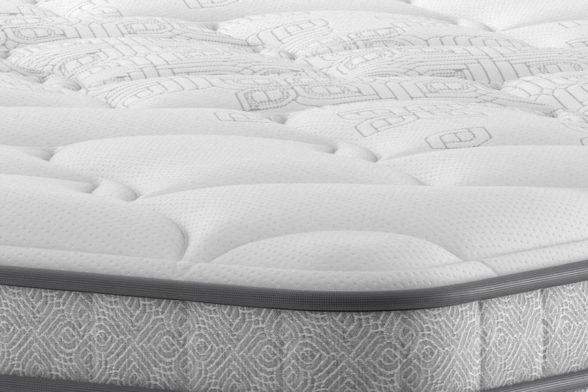A full shot of the Spencer Plush Mattress, shown on a dark blue divan base with a tall, contemporary upholstered headboard, set in a bright bedroom with light wood wall panelling. The mattress has a deep quilted white top.
